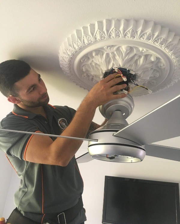 Electrician installing a ceiling fan and connecting its wires to an ornate ceiling medallion, wearing a grey uniform.