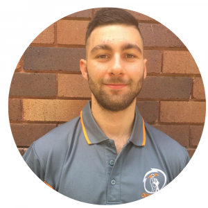 Young man with short dark hair and a beard, wearing a gray collared shirt with a logo, standing against a brick wall.