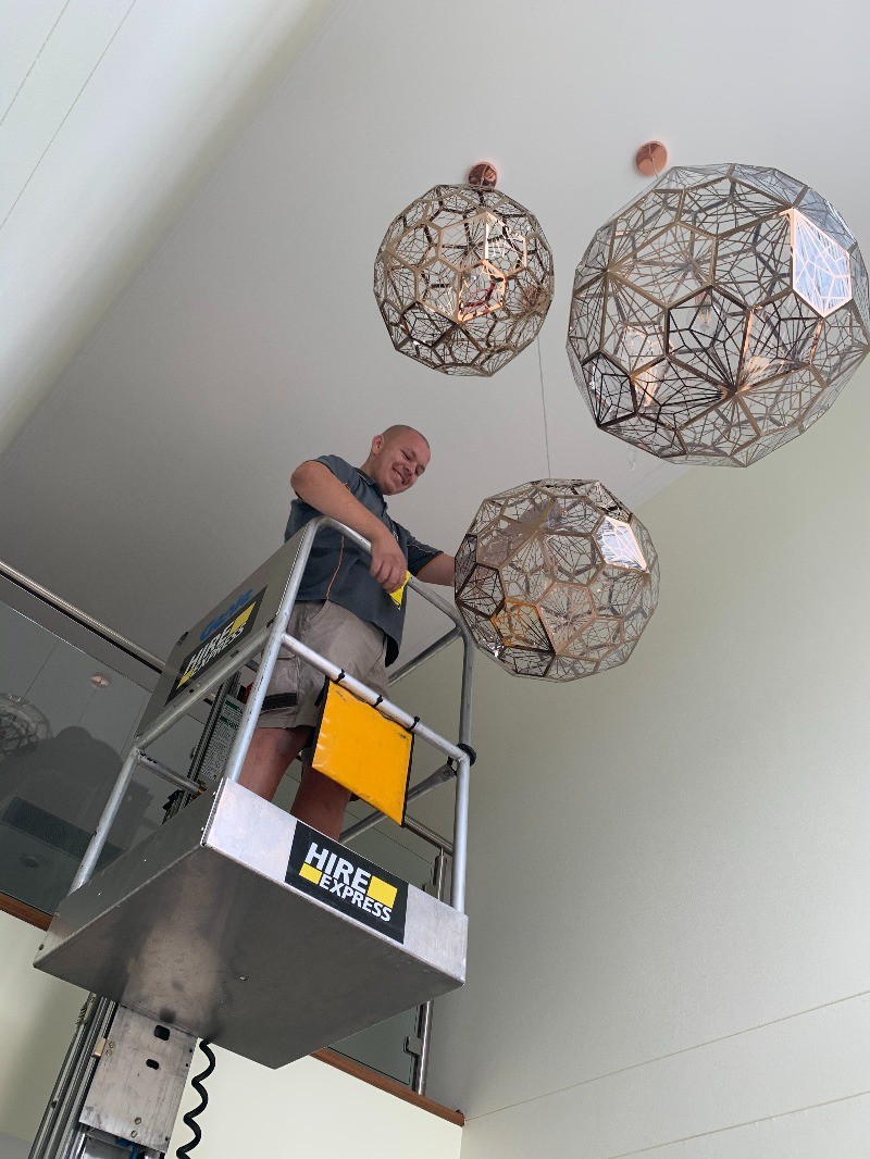 A man standing on a lift repairing or installing geometric pendant lights in a high-ceilinged room.