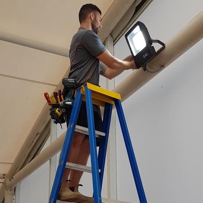 A man stands on a blue ladder fixing a light fixture, with tools attached to his belt, inside an industrial building.