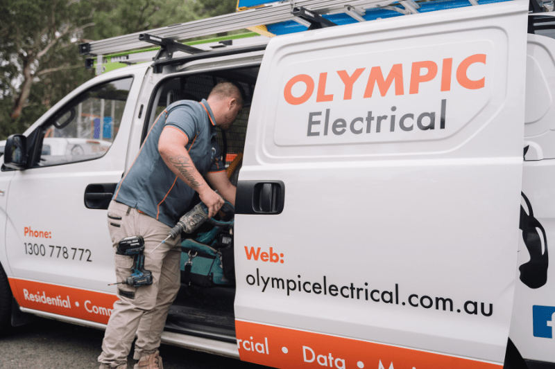 Electrician accessing tools from the back of an Olympic Electrical van with company information displayed on the side.
