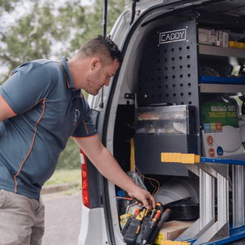 Untitled design (26) A man organizing tools in the back of a work van equipped with shelves, bins, and ladders, wearing a navy shirt and light shorts.