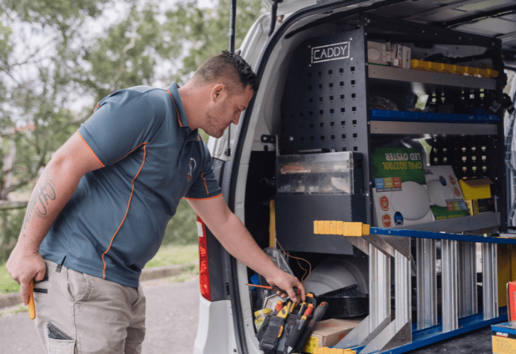 A man organizing tools in the back of a work van equipped with shelves, bins, and ladders, wearing a navy shirt and light shorts.