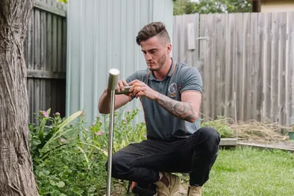 A man kneels in a backyard garden, inspecting a soil sensor with a screwdriver near a fence and storage shed.