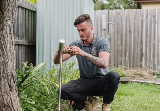 A man kneels in a backyard garden, inspecting a soil sensor with a screwdriver near a fence and storage shed.