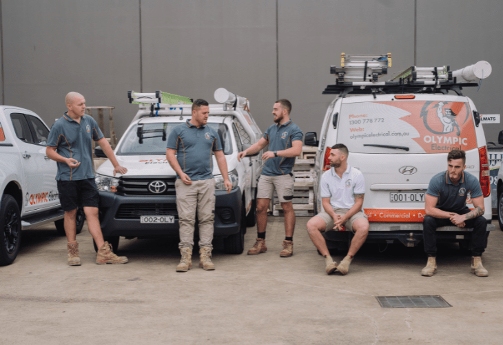 Five men in work attire stand and sit in front of three branded electrical service vehicles in an outdoor area.