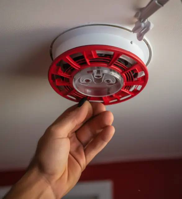 Hand reaching towards a ceiling-mounted smoke detector with a red cover, likely for maintenance or testing.