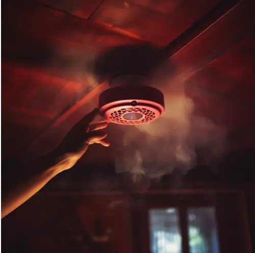 Hand reaching up to a smoke detector on the ceiling with smoke surrounding it.