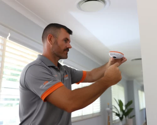 A man in a gray polo shirt installs a smoke detector on a white ceiling in a bright room with large windows.