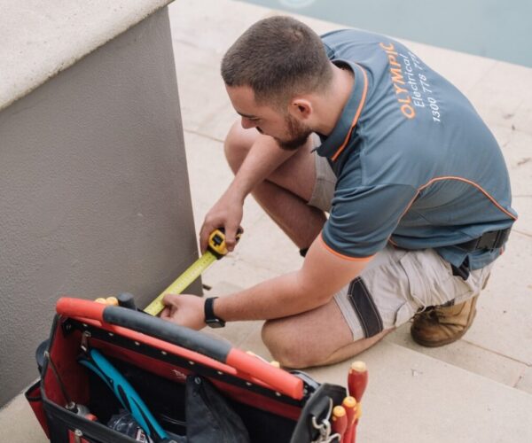 Technician kneeling and measuring next to a pool with a tape measure. Toolbag with various tools rests on the steps beside him.