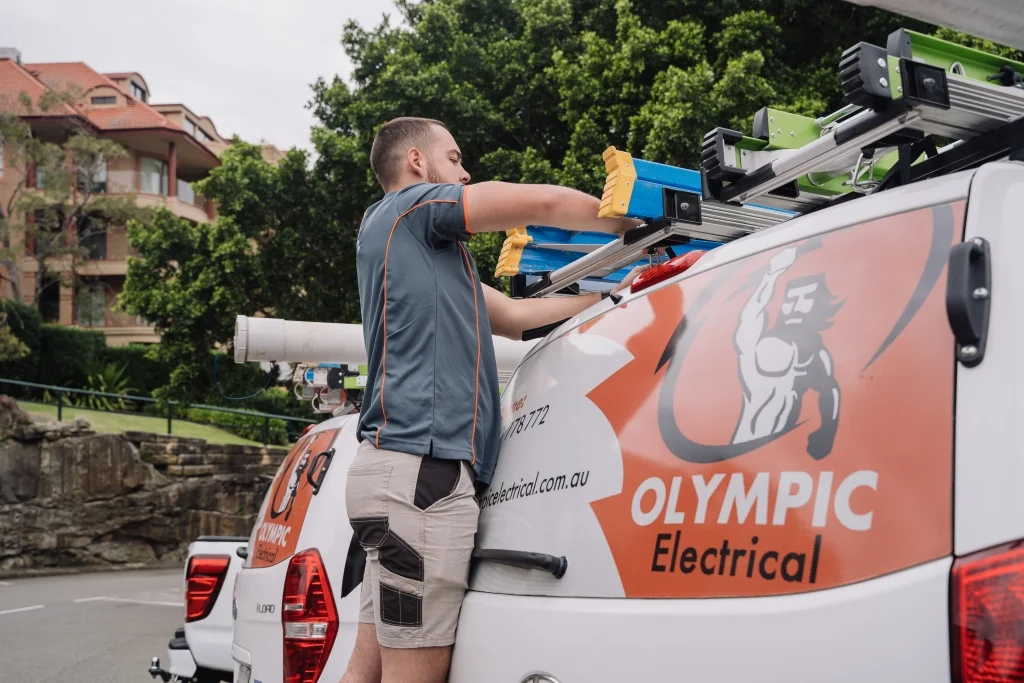 An electrician retrieves tools from a van with Olympic Electrical branding, parked in a residential area.