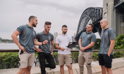 Five men in uniforms discuss something on a tablet near a large steel bridge and lush greenery in the background.