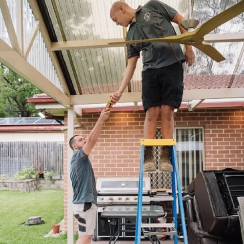 Two men in work uniforms, one on a ladder, installing a ceiling fan under a patio roof in a backyard setting.