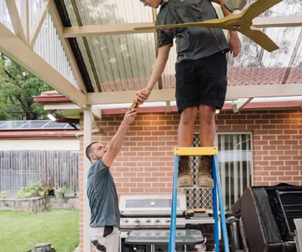 Two men in work uniforms, one on a ladder, installing a ceiling fan under a patio roof in a backyard setting.
