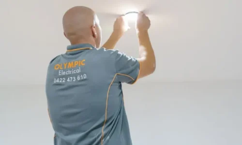 Electrician from Olympic Electrical installing a ceiling light fixture while wearing a gray company uniform.