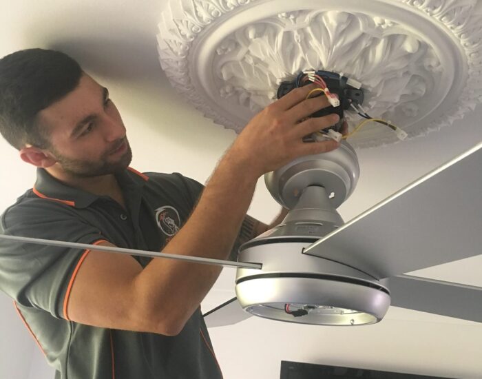 A technician in a grey shirt uses tools to install a wooden ceiling fan in a room