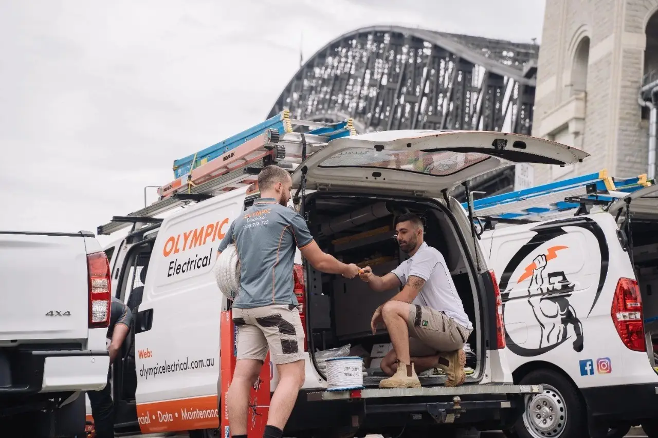 Two electricians are working outside a van loaded with equipment near a bridge, with one inside the vehicle and the other standing.