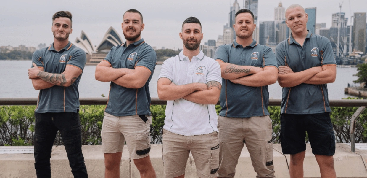 Five men in uniform pose with arms crossed, standing in front of the Sydney Opera House and city skyline.